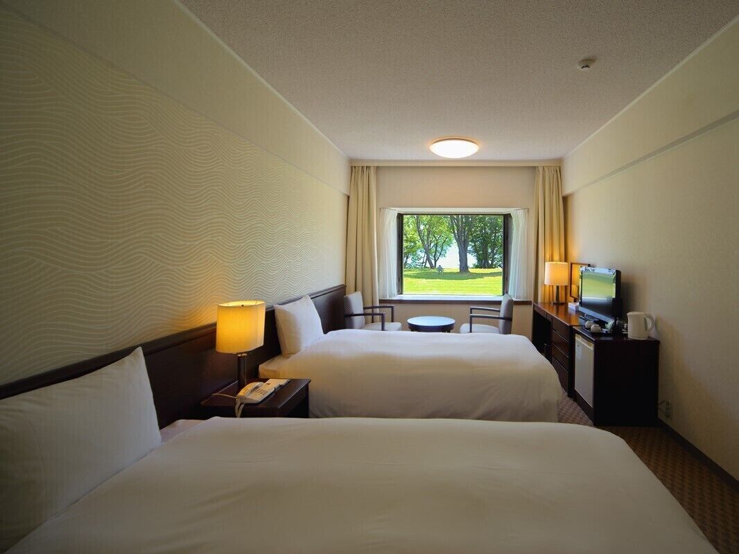 A hotel room with two beds, a television, a desk with a lamp, and a view of a grassy area through a window.