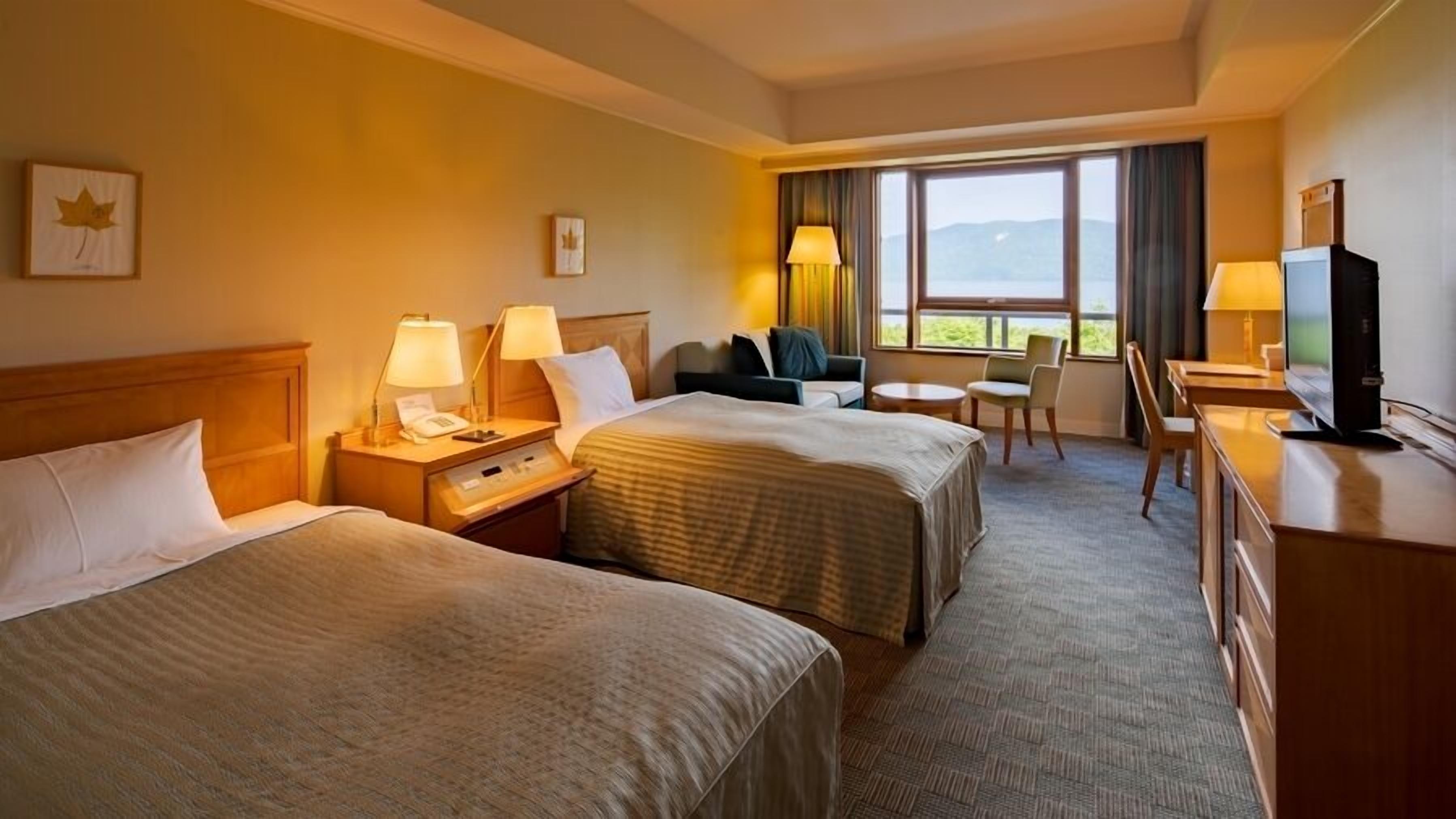 A hotel room with two beds, a desk with a TV, a chair, and a view of the outdoors.