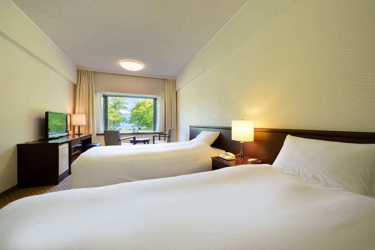 A hotel room with two beds, a desk with a TV, a lamp, and a window with curtains.