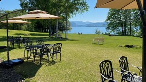 Un patio al aire libre con mesas y sillas, rodeado de árboles y con vista a un lago con montañas a lo lejos.