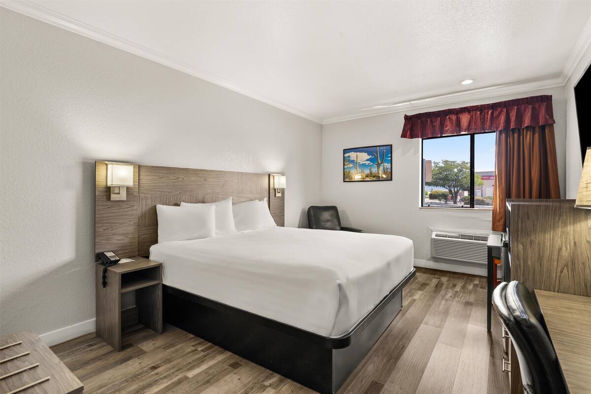 A hotel room with a large bed, a bedside table with a lamp, a chair, a desk, and a window with curtains.
