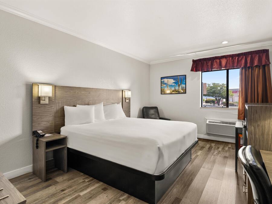 Habitación de hotel con una cama grande, una mesita de noche con lámpara, una silla, un escritorio y una ventana con cortinas.
