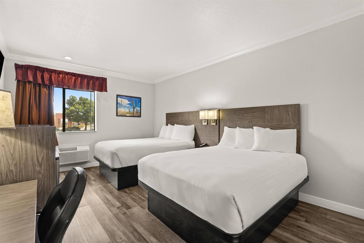 A hotel room with two beds, a desk, a chair, a window with curtains, and a painting on the wall.