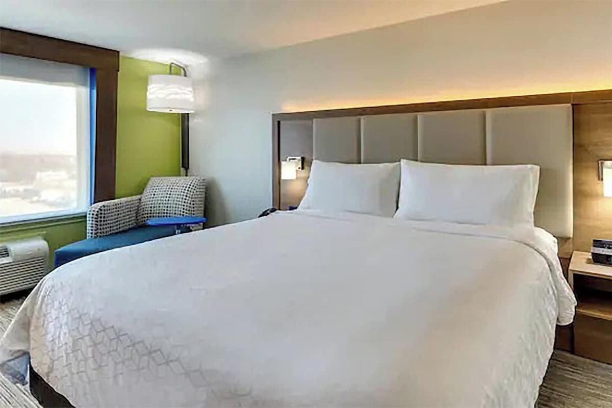 Uma cama grande com cabeceira acolchoada, uma mesinha de cabeceira com rádio e uma cadeira com almofada estampada.