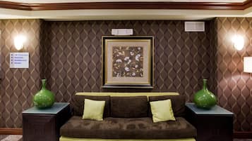 A hotel room with a brown patterned wall, a framed floral artwork, a brown sofa with cushions, two green vases, and a sign indicating guest rooms and amenities.