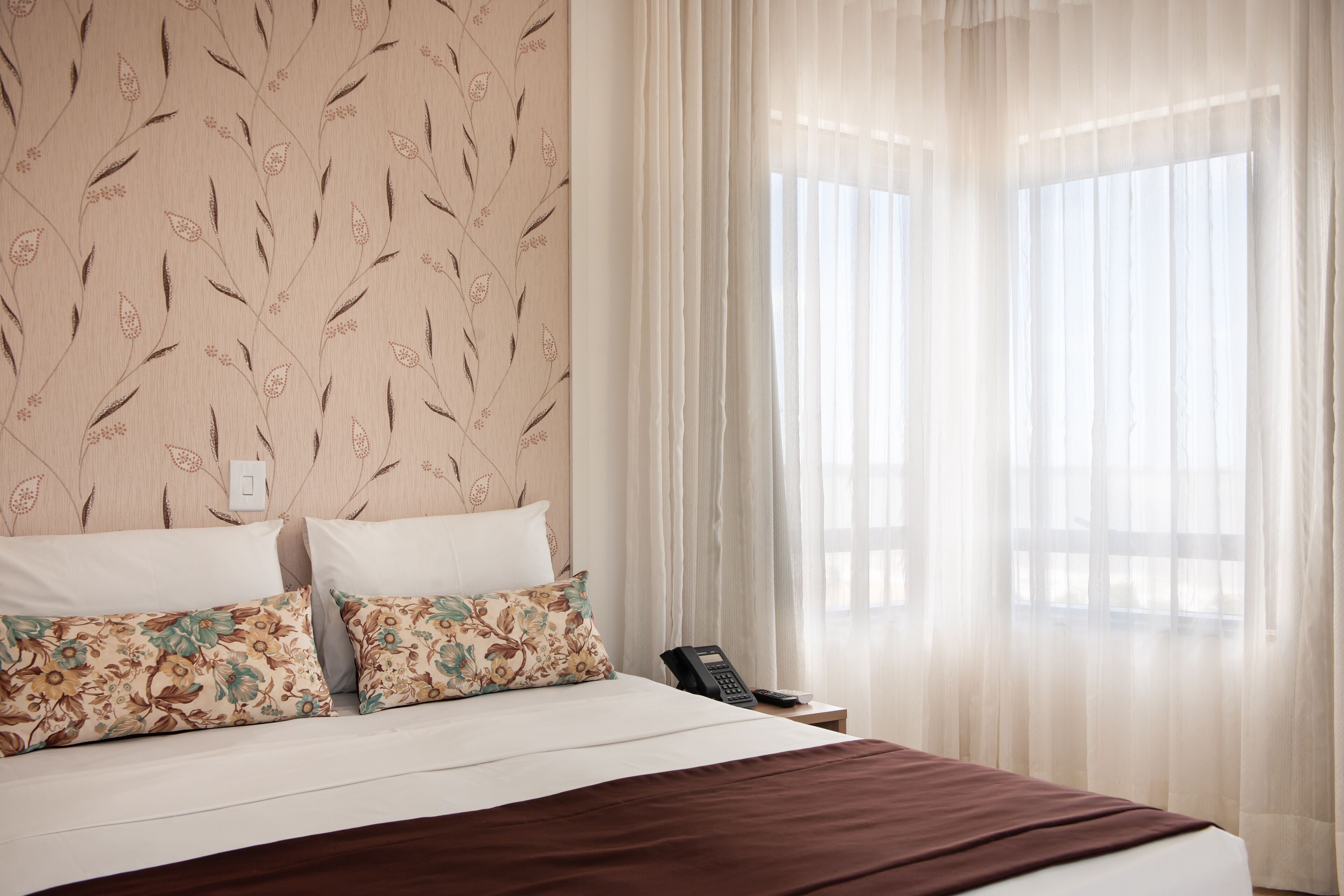 A hotel room with a bed, two pillows, a bedside table with a phone, and sheer curtains.