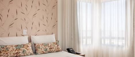 A hotel room with a bed, two pillows, a bedside table with a phone, and sheer curtains.
