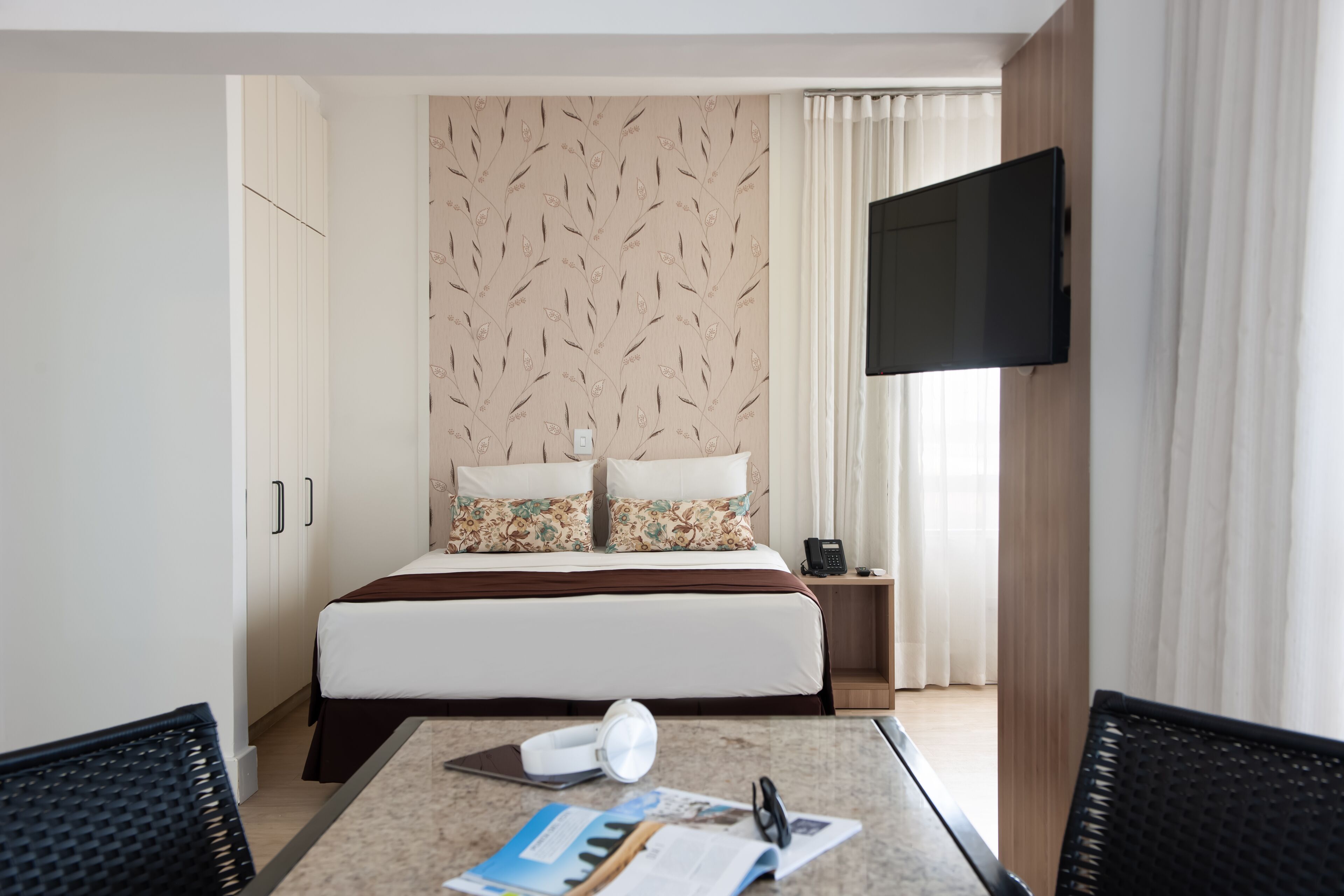 A hotel room with a bed, a desk with a chair, a TV, and a window with curtains.
