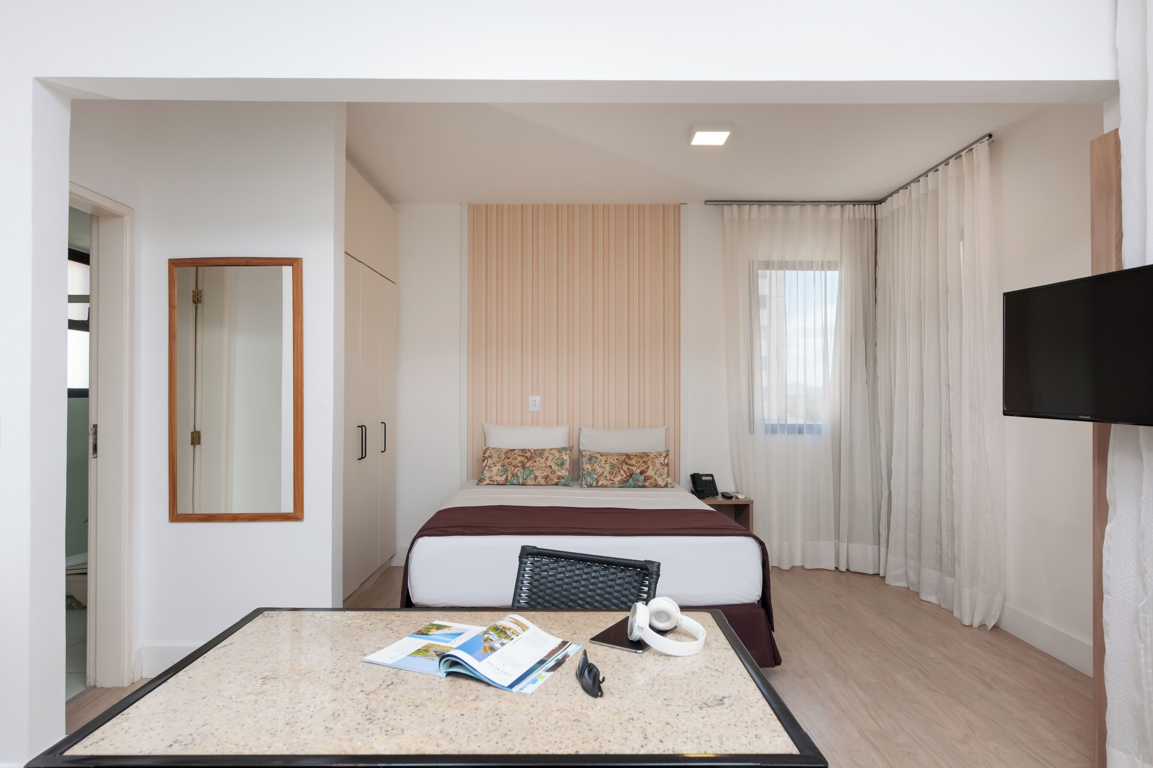 Quarto de hotel com cama, televisão, secretária com revista e janela com cortinas.
