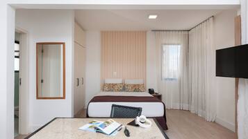 Quarto de hotel com cama, televisão, secretária com revista e janela com cortinas.