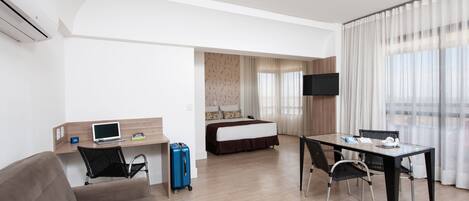 Habitación de hotel con cama, escritorio con computadora portátil, televisión y zona de comedor con sillas.