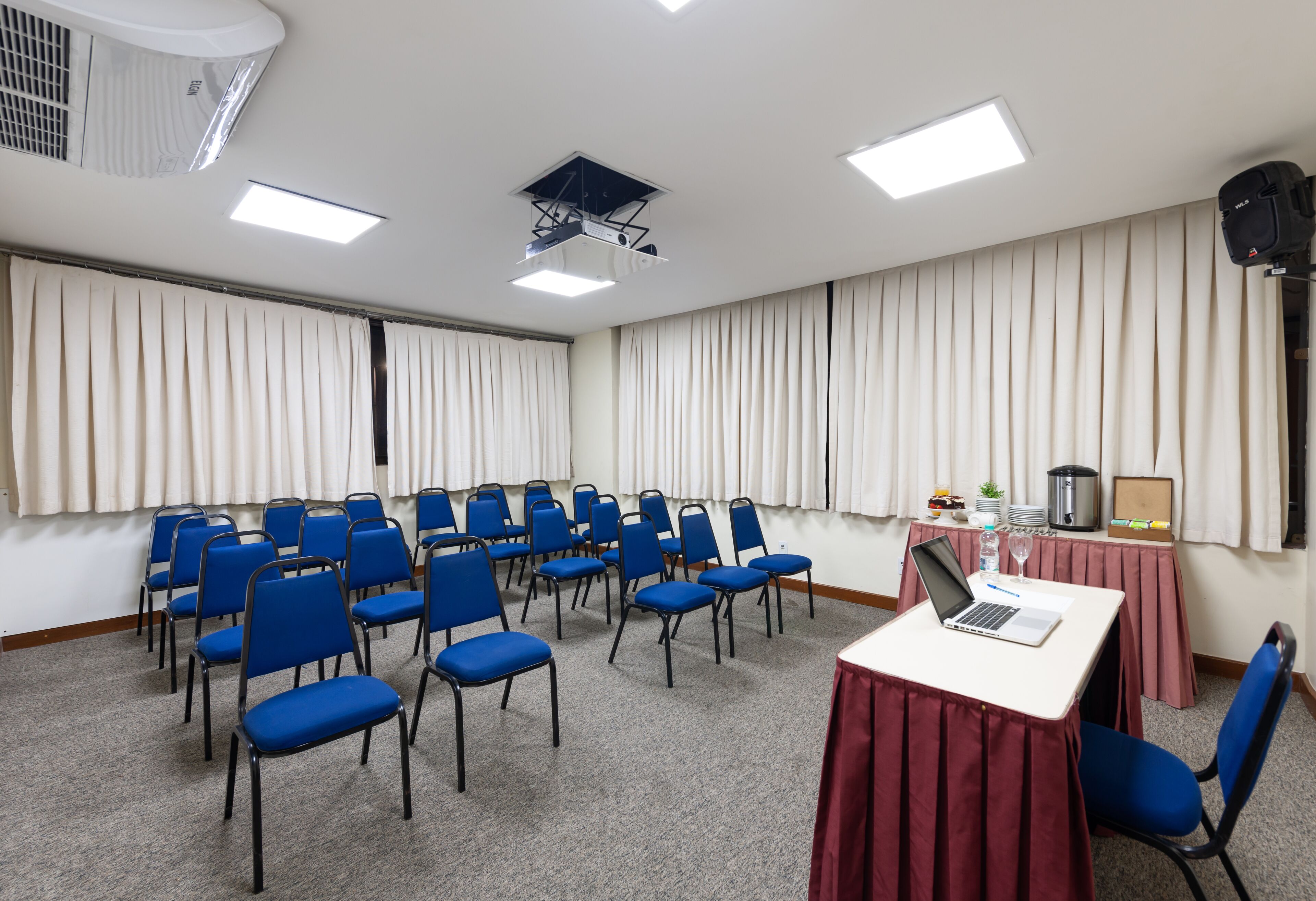 A conference room with rows of blue chairs, a white table with a laptop and a glass of water, and a projector on the ceiling.