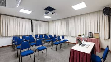 Uma sala de conferências com fileiras de cadeiras azuis, uma mesa branca com um laptop e um copo de água, e um projetor no teto.