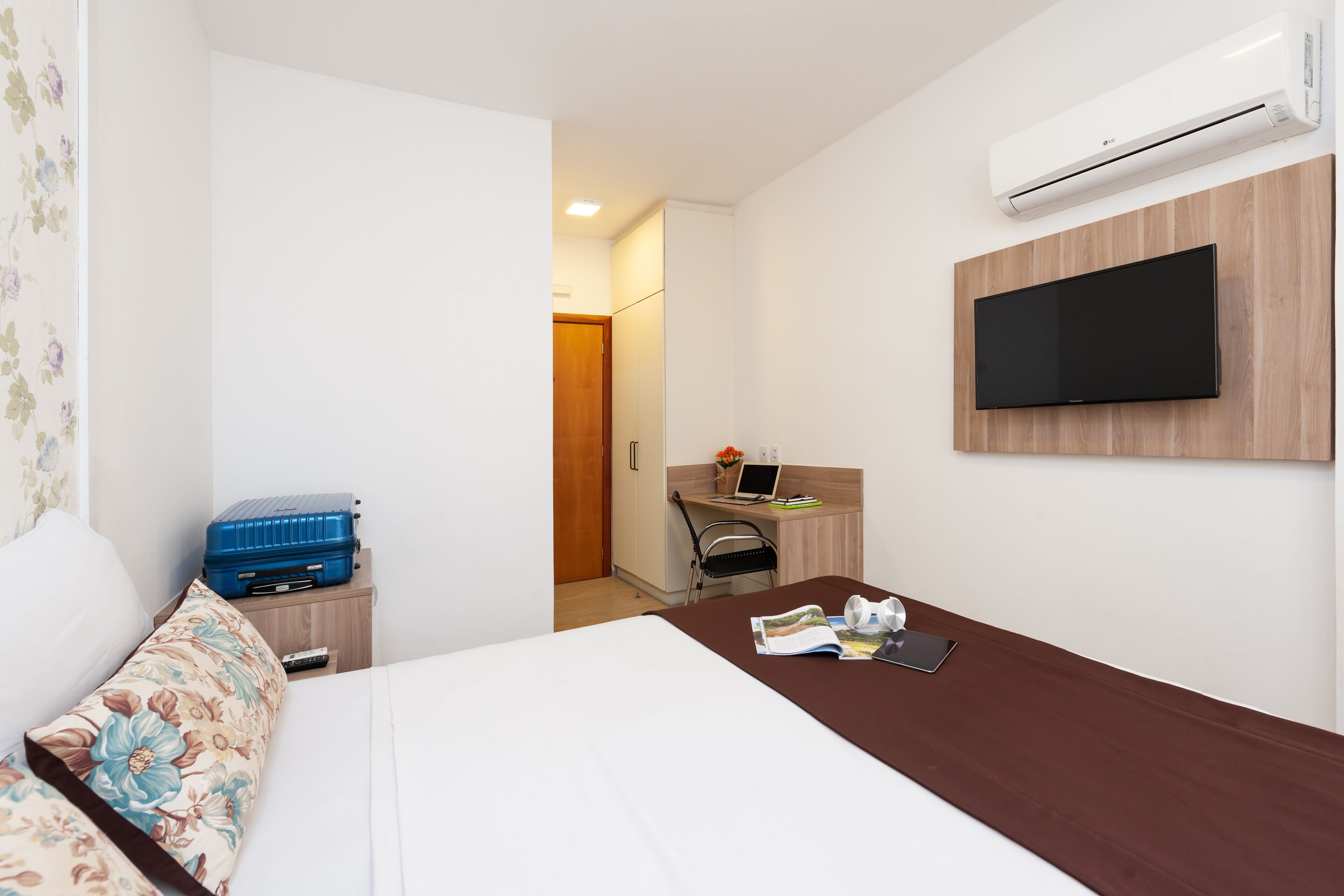 Een hotelkamer met een bed, een bureau met een laptop, een aan de muur bevestigde televisie en een airconditioning.