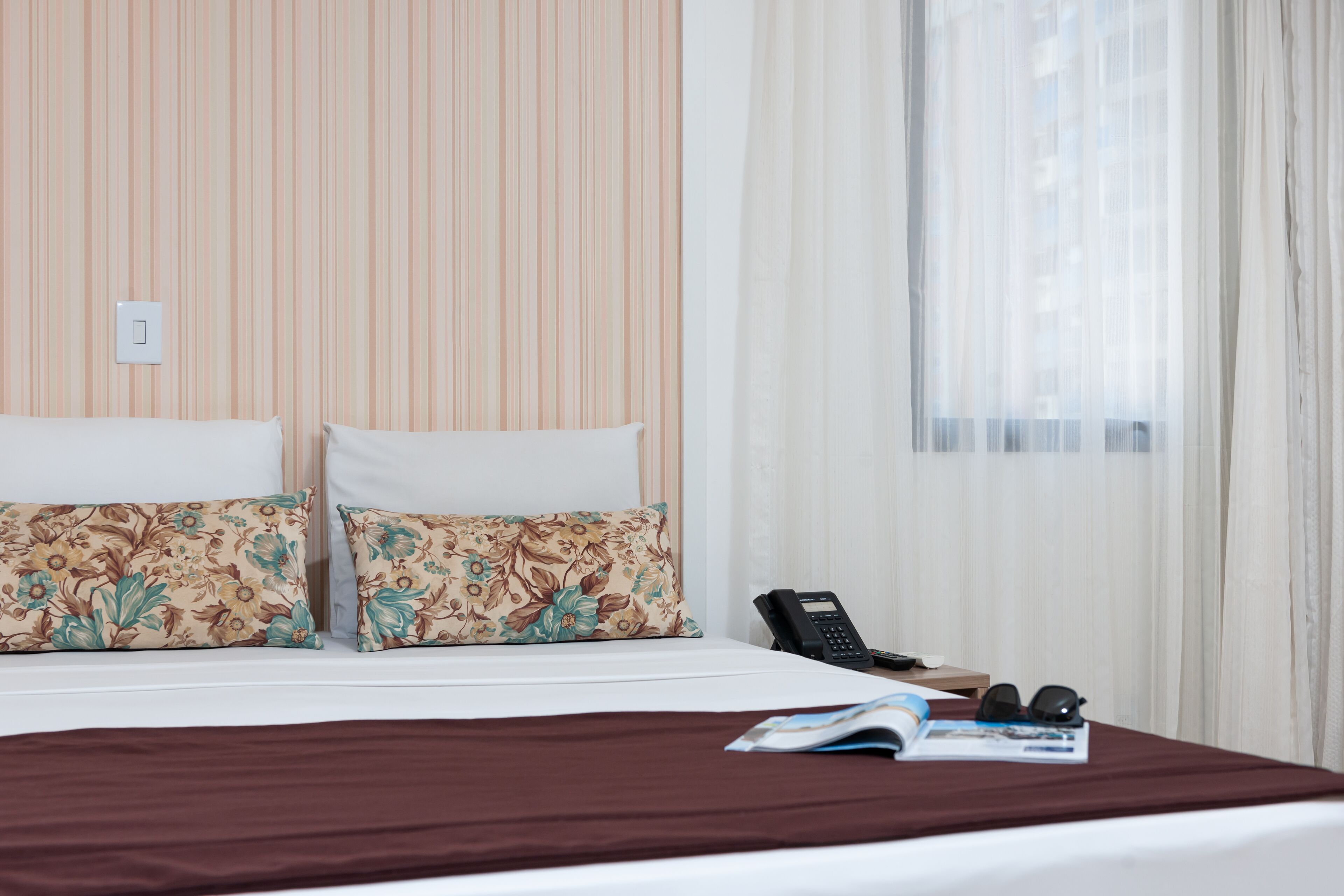 A hotel room with a bed, two floral-patterned pillows, a brown bedspread, a telephone, and a magazine on the bed.
