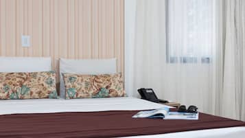 Quarto de hotel com uma cama, duas almofadas com padrão floral, um espalha cama marrom, um telefone e uma revista sobre a cama.