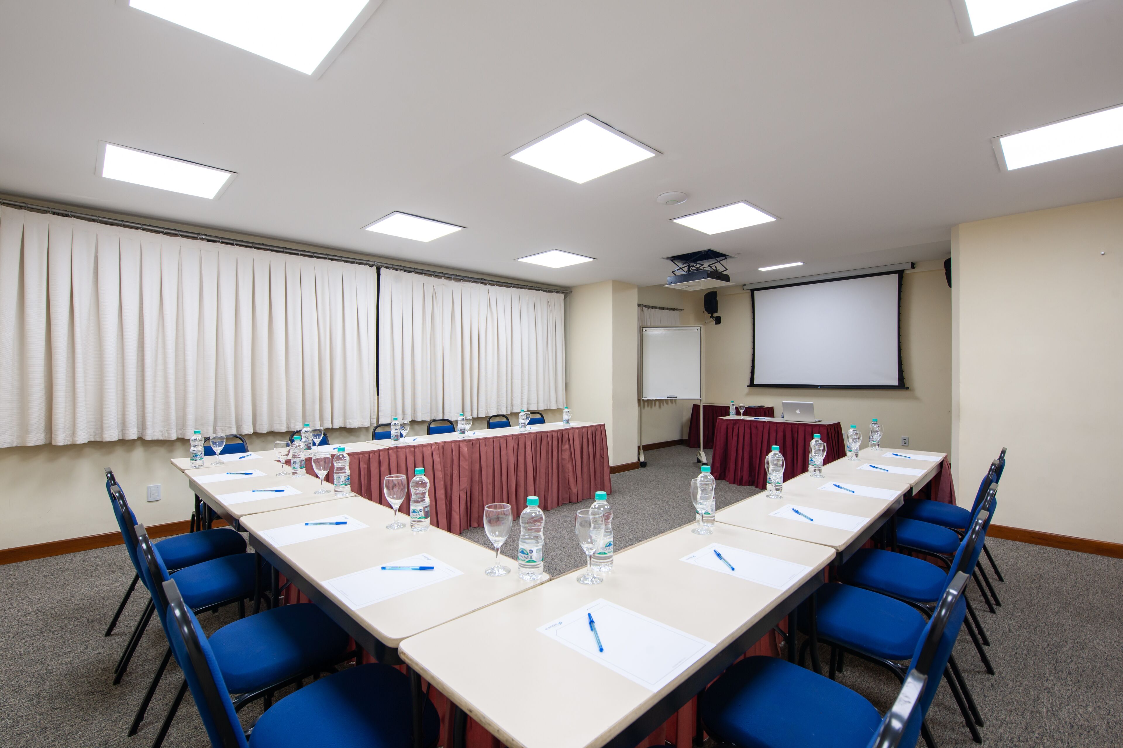 Un salón de conferencias con una larga mesa, sillas azules, botellas de agua y una pantalla de proyector.