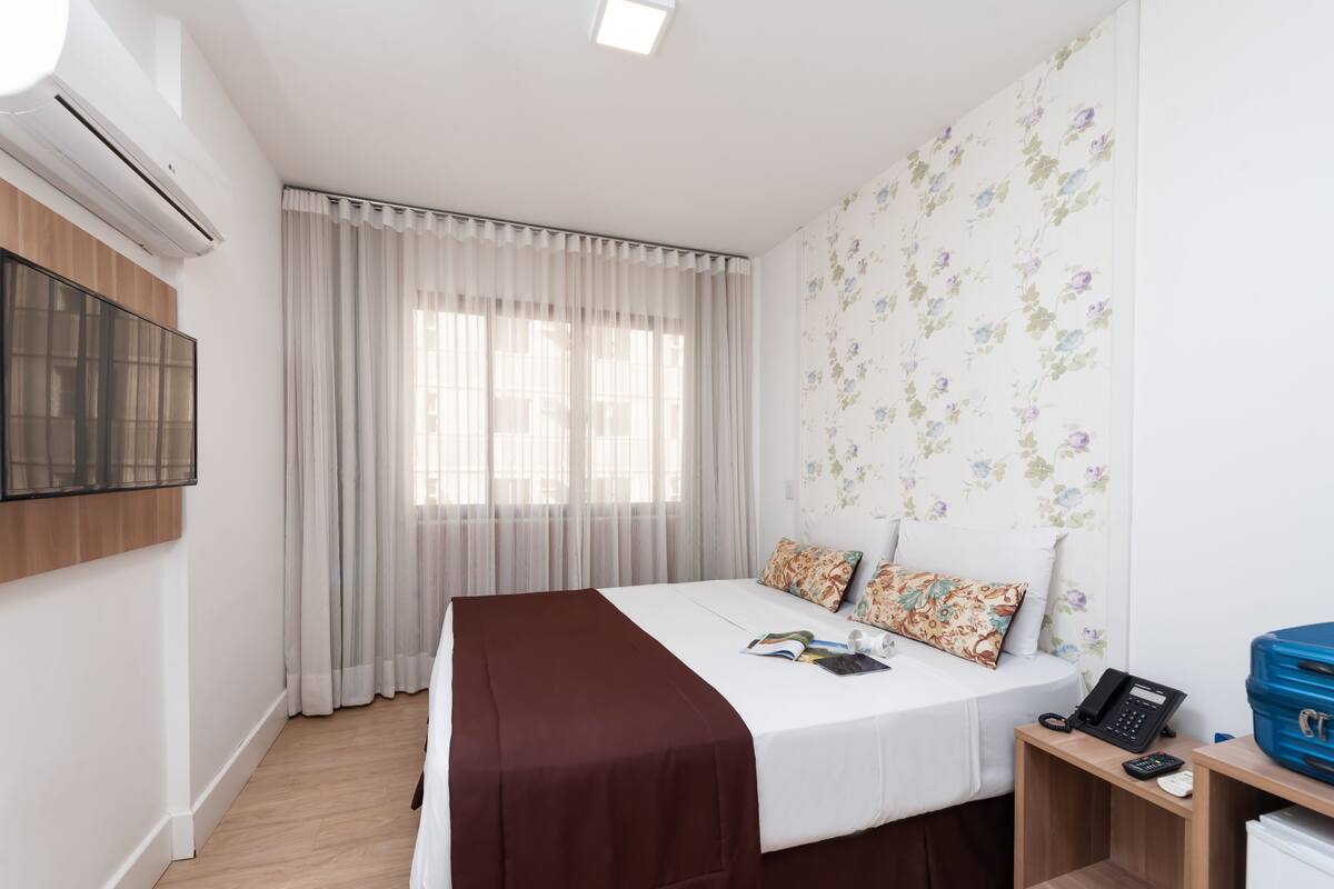 Ein Hotelzimmer mit einem Bett, einem Nachttisch mit Telefon und Fernbedienung, einem blauen Koffer und einem Fenster mit Vorhängen.