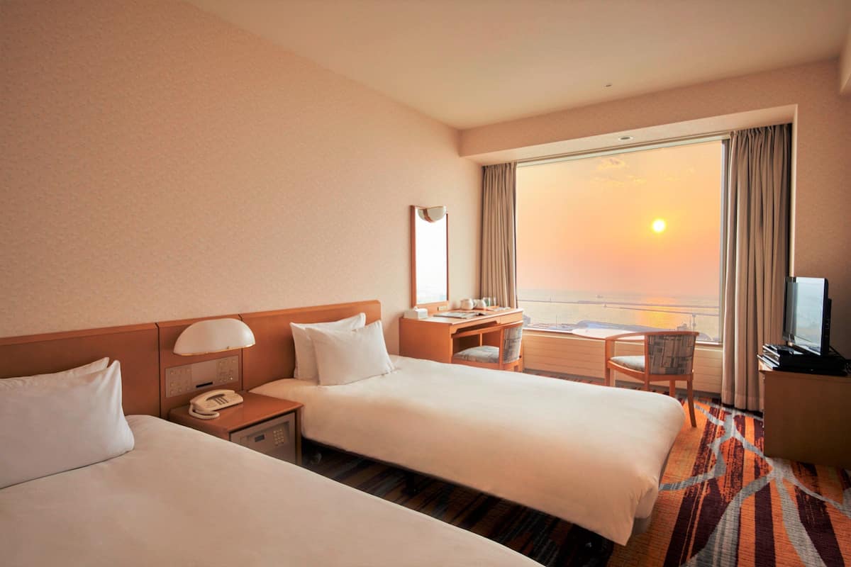 A hotel room with two beds, a desk, a chair, a TV, and a large window with a view of the sunset.