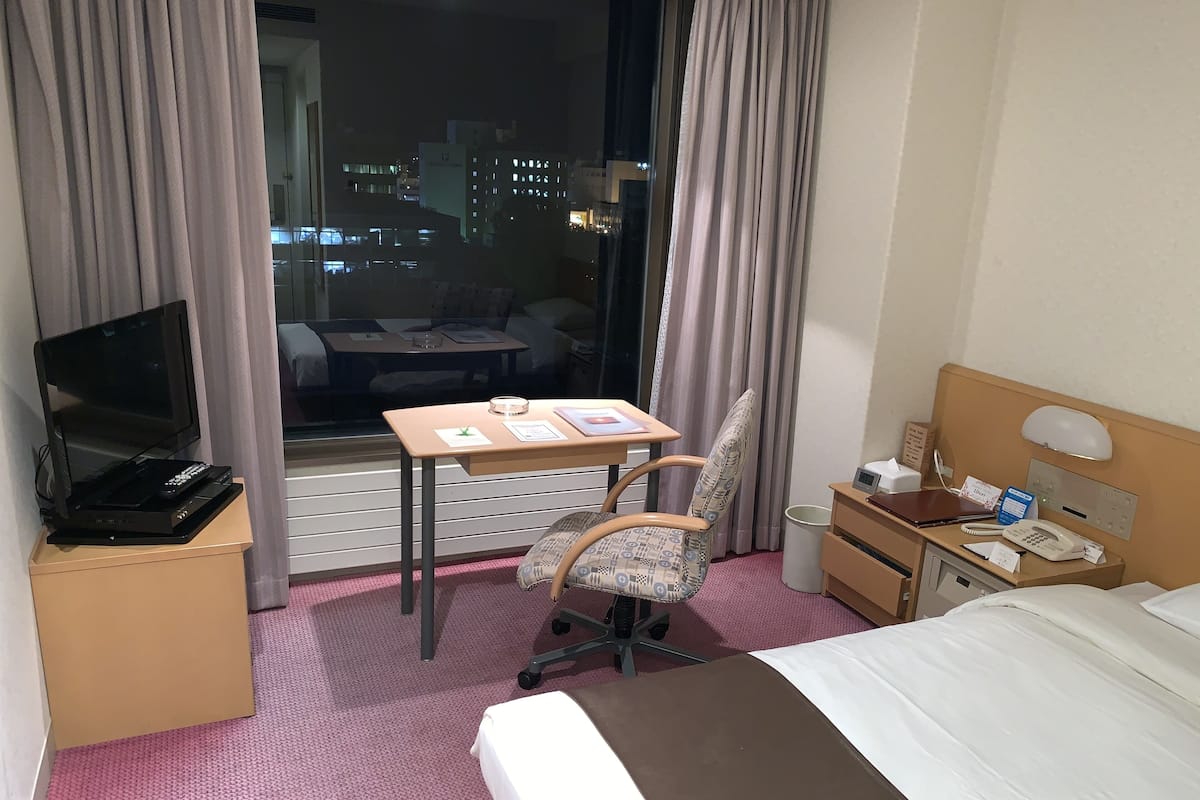 A hotel room with a bed, a desk with a chair, a TV, a phone, and a window with curtains.