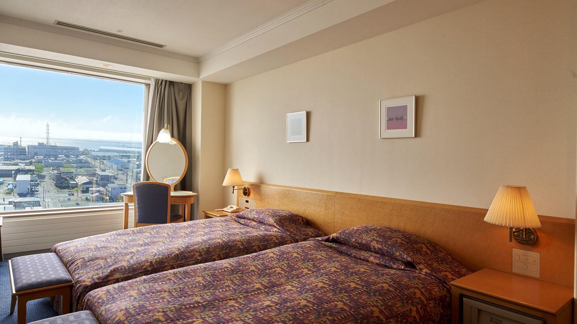 A hotel room with two beds, a desk, a chair, a mirror, two wall-mounted lamps, and a large window offering a city view.