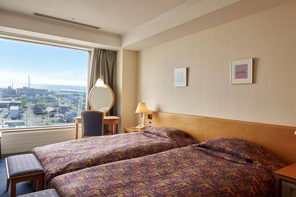 A hotel room with two beds, a desk, a chair, a mirror, two wall-mounted lamps, and a large window offering a city view.