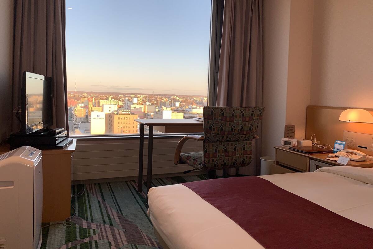 A hotel room with a bed, a desk with a television, a chair, a small table, a lamp, and a view of the cityscape.