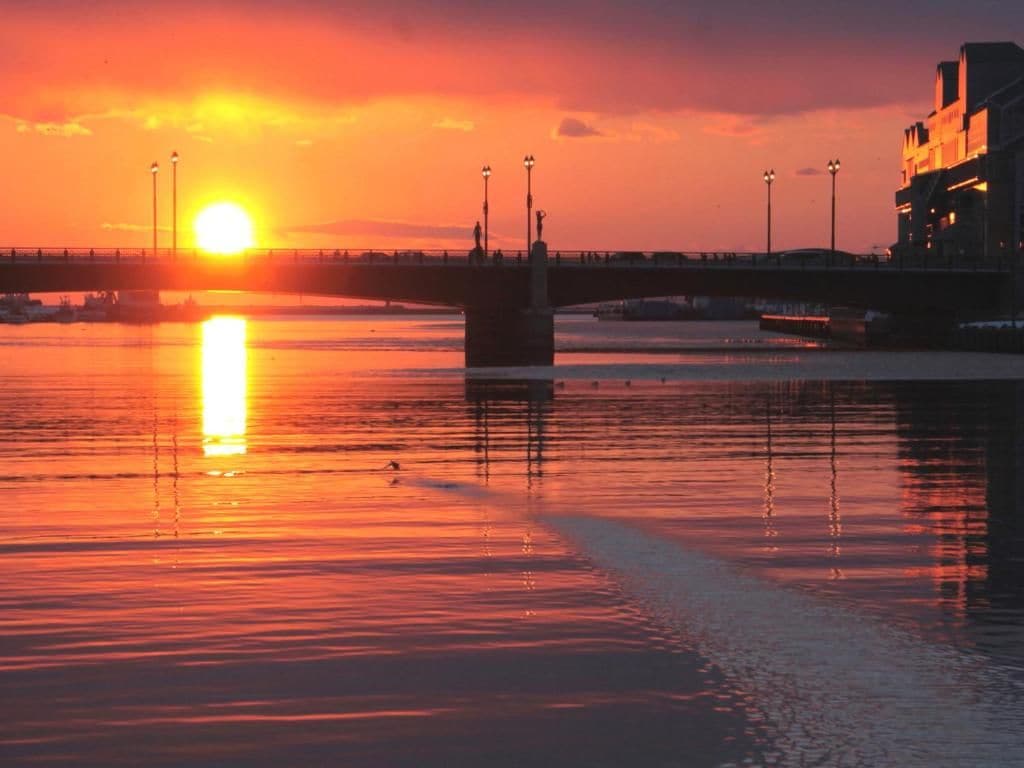 Un atardecer sobre un río, con un puente y edificios al fondo.
