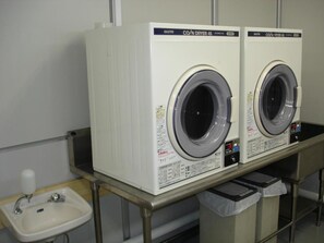 Two identical front-loading washing machines in a commercial setting, with a sink and a trash bin nearby. - Kushiro Prince Hotel (Kushiro)