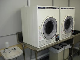 Two identical front-loading washing machines in a commercial setting, with a sink and a trash bin nearby.