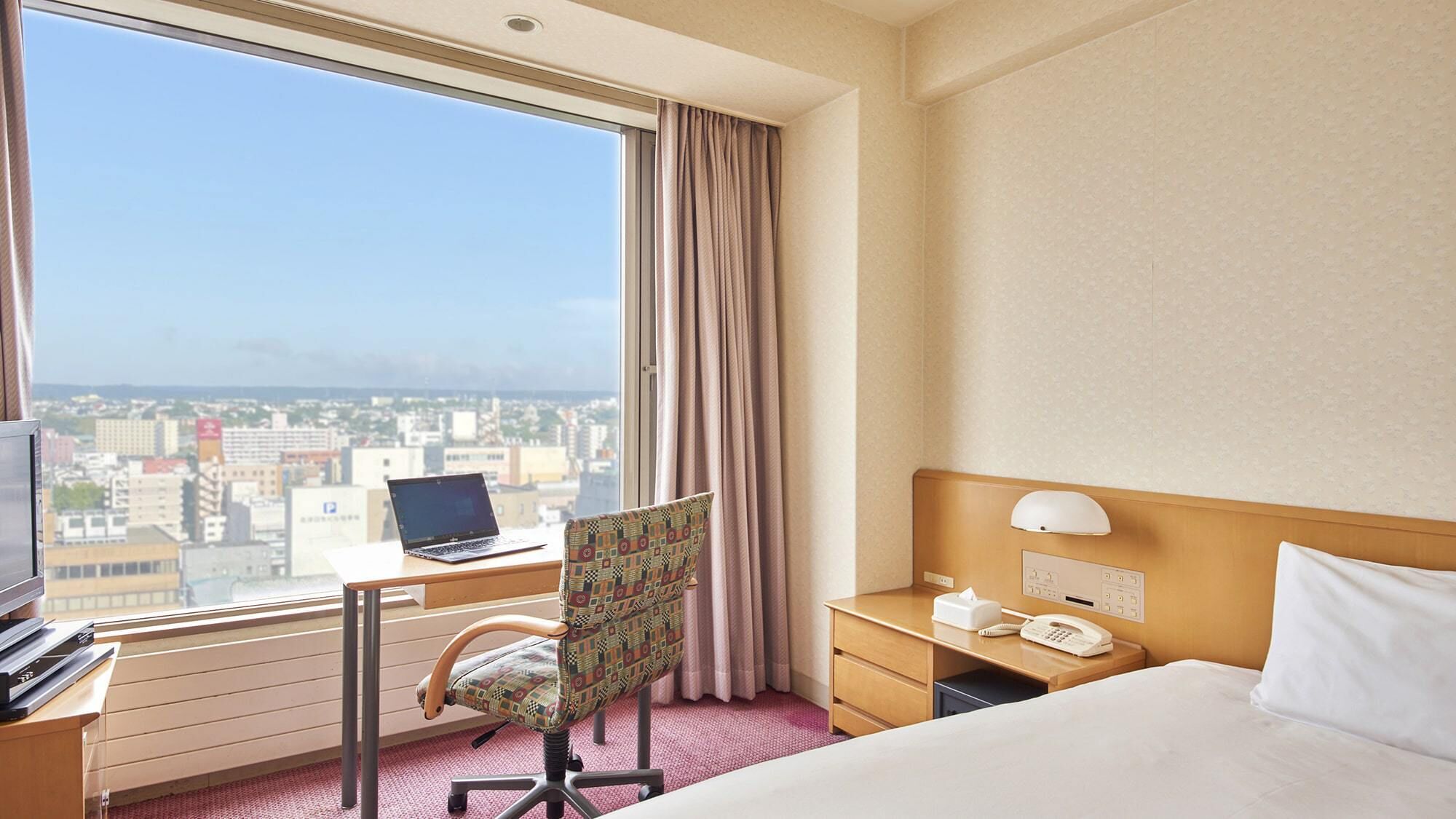 A hotel room with a bed, a desk with a laptop, a chair, a bedside table with a phone, and a window with a city view.