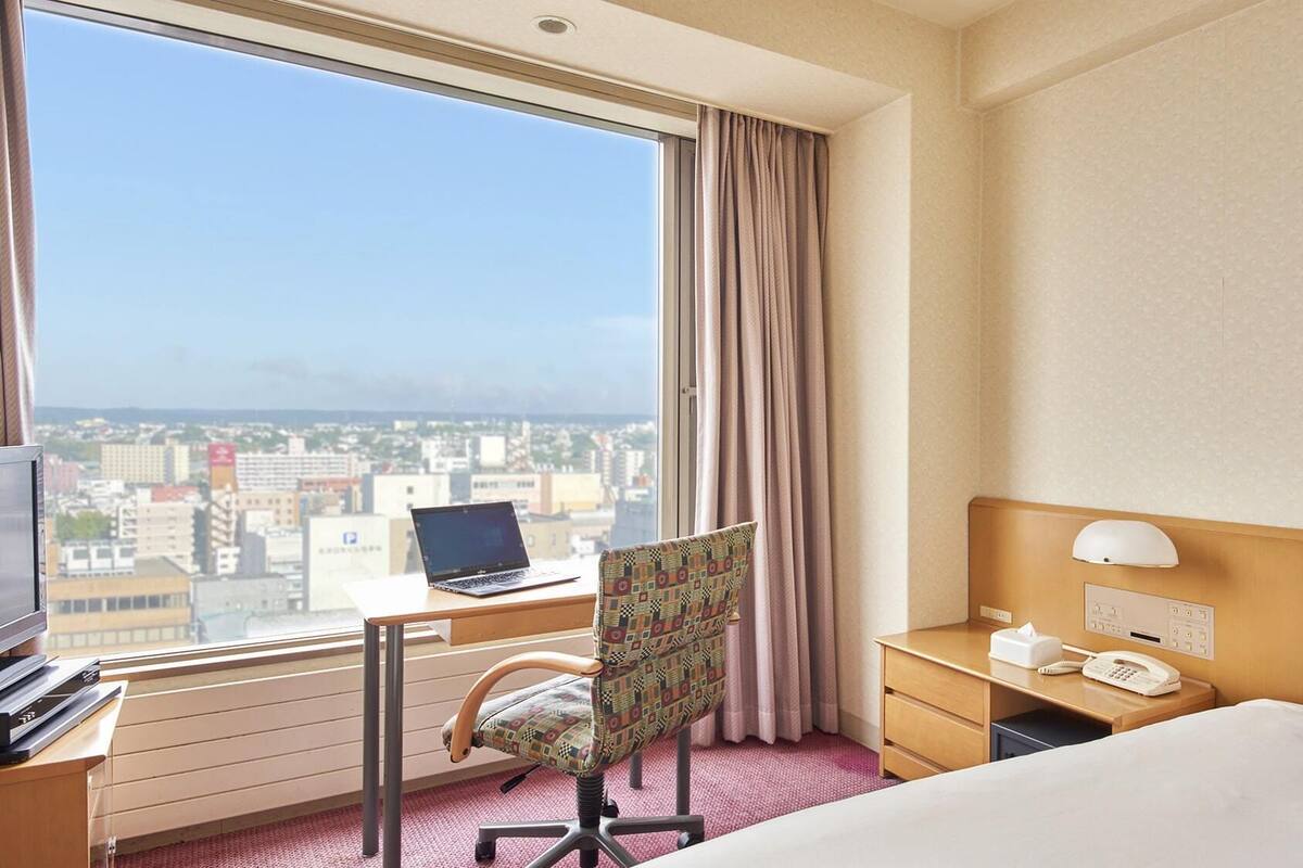 A hotel room with a bed, a desk with a laptop, a chair, a bedside table with a phone, and a window with a city view.