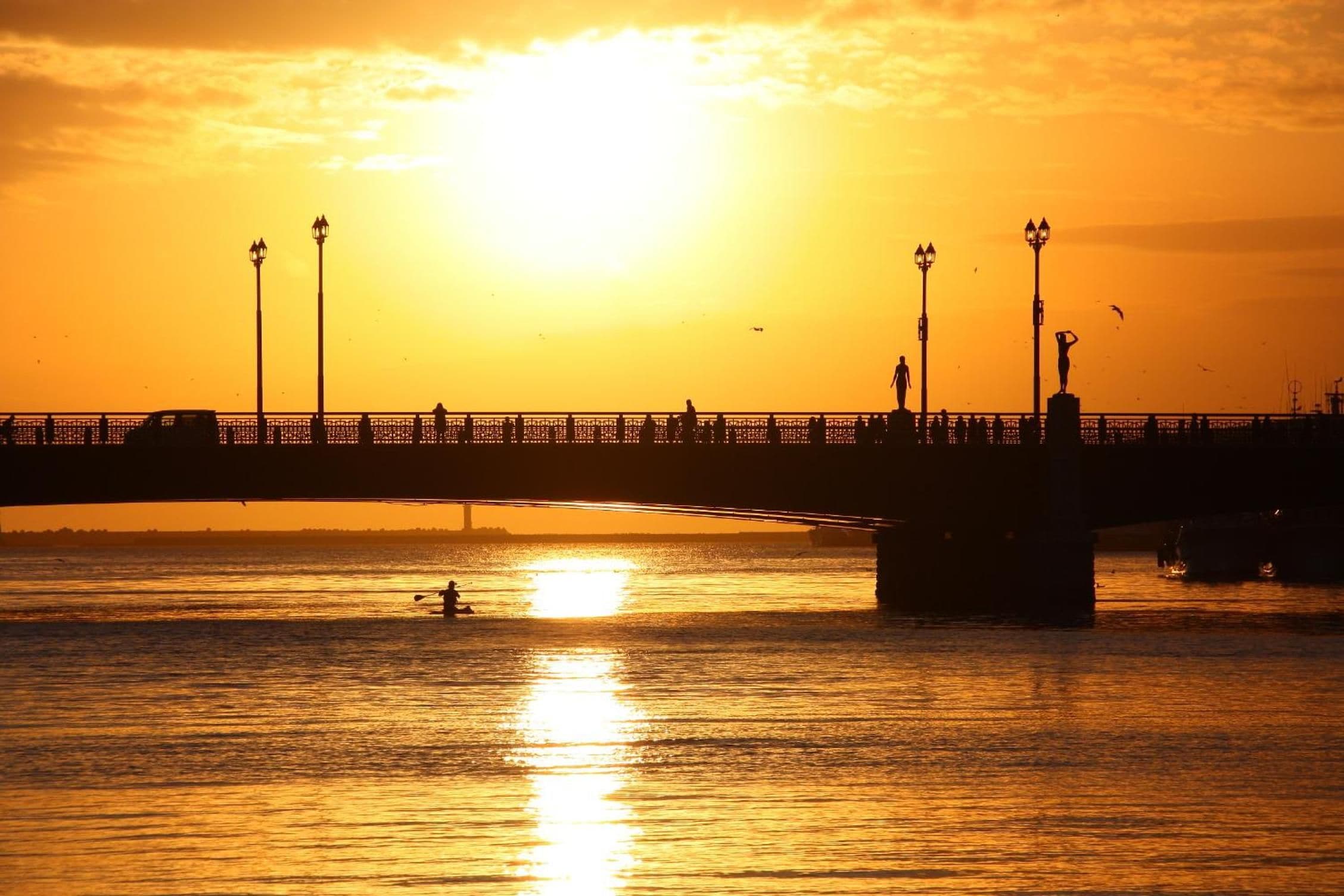 A sunset over a waterfront with a bridge, street lamps, and a person kayaking.