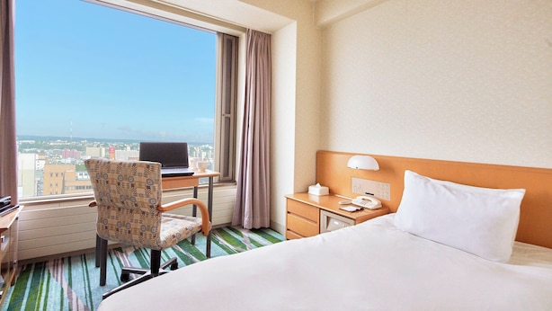 A hotel room with a large bed, a desk with a computer, a chair, a nightstand, and a view of the city through the window. - Kushiro Prince Hotel (Kushiro)