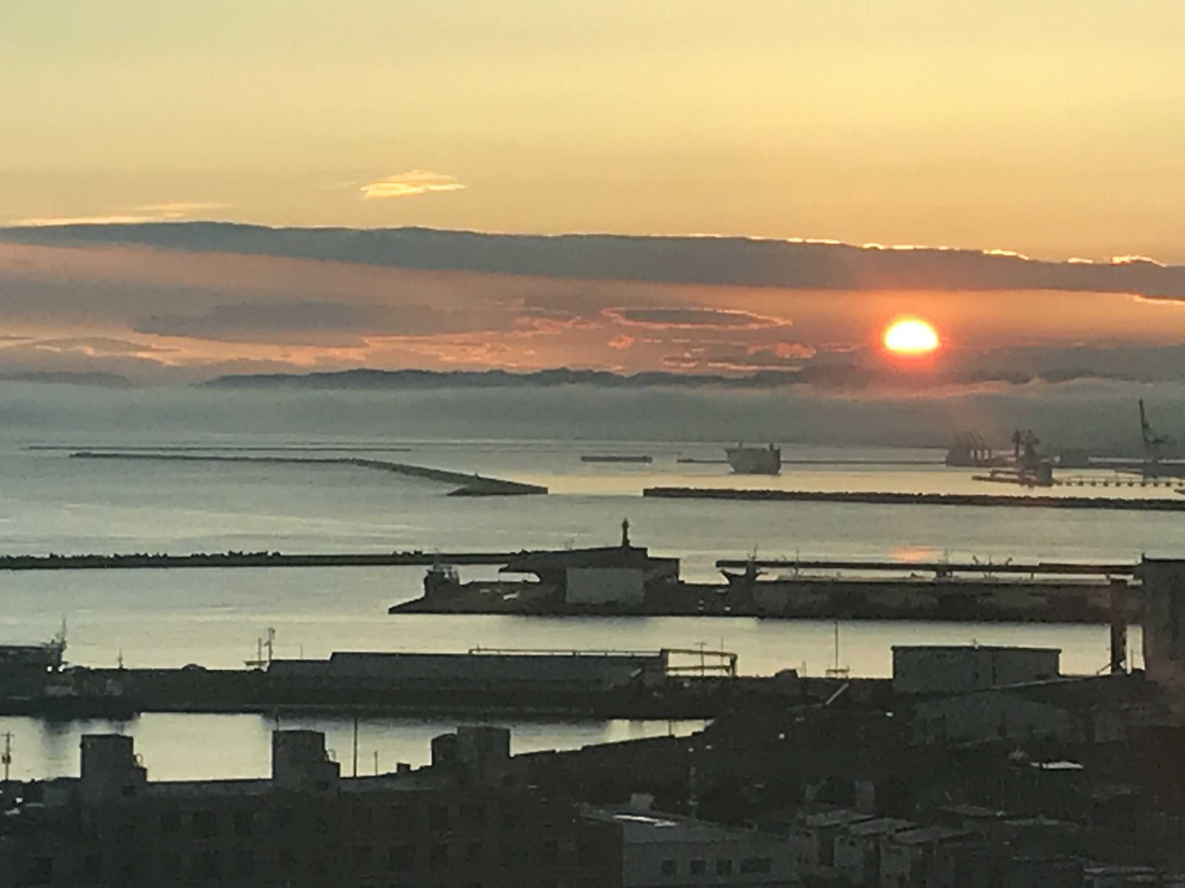A coastal cityscape during sunset with buildings and boats.
