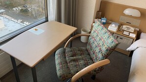 A hotel room with a bed, a desk, a chair, a bedside table with a phone and tissues, and a window with a view of a parking lot and buildings. - Kushiro Prince Hotel (Kushiro)