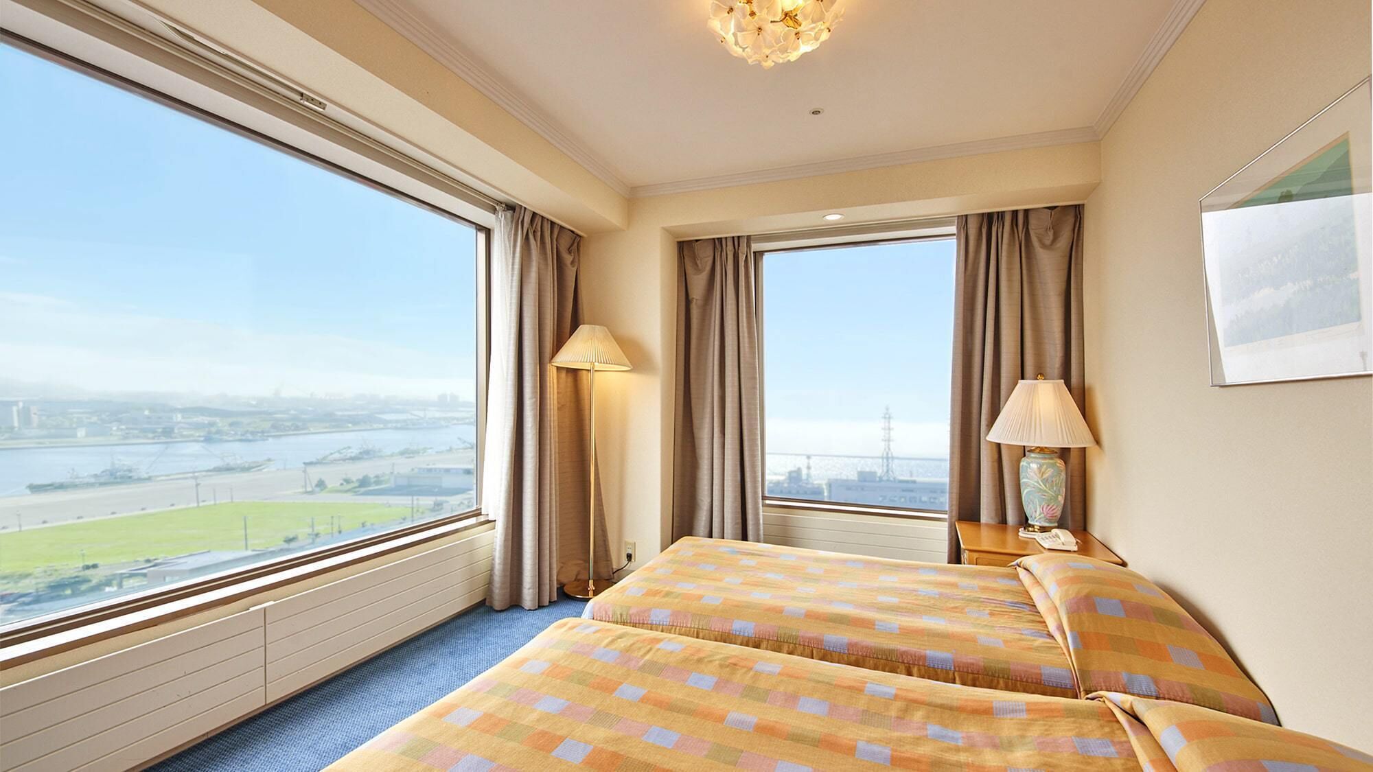 A hotel room with two beds, large windows, a view of a cityscape, and a lamp on the nightstand.