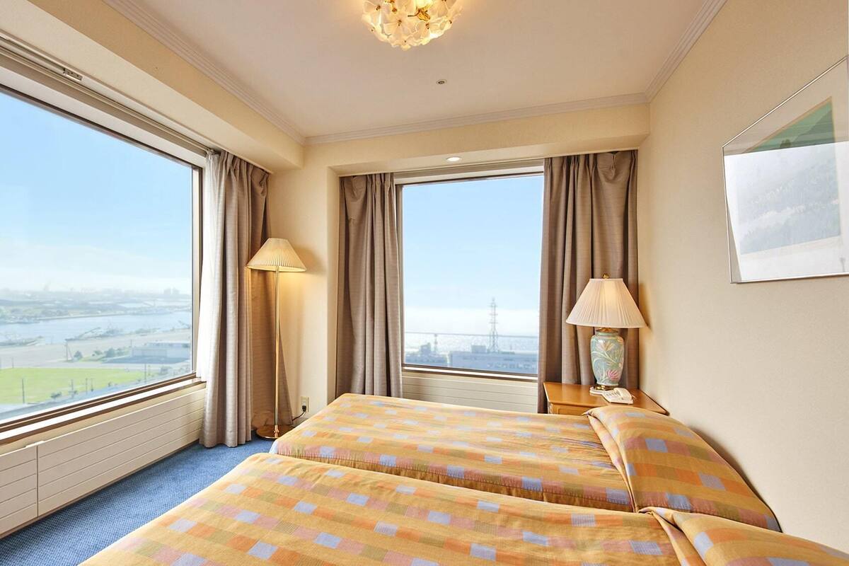 A hotel room with two beds, large windows, a view of a cityscape, and a lamp on the nightstand.