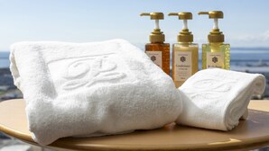 A white towel with a monogram, two bottles of conditioner, and a bottle of body soap on a round table with a clear view of the sky in the background. - Kushiro Prince Hotel (Kushiro)