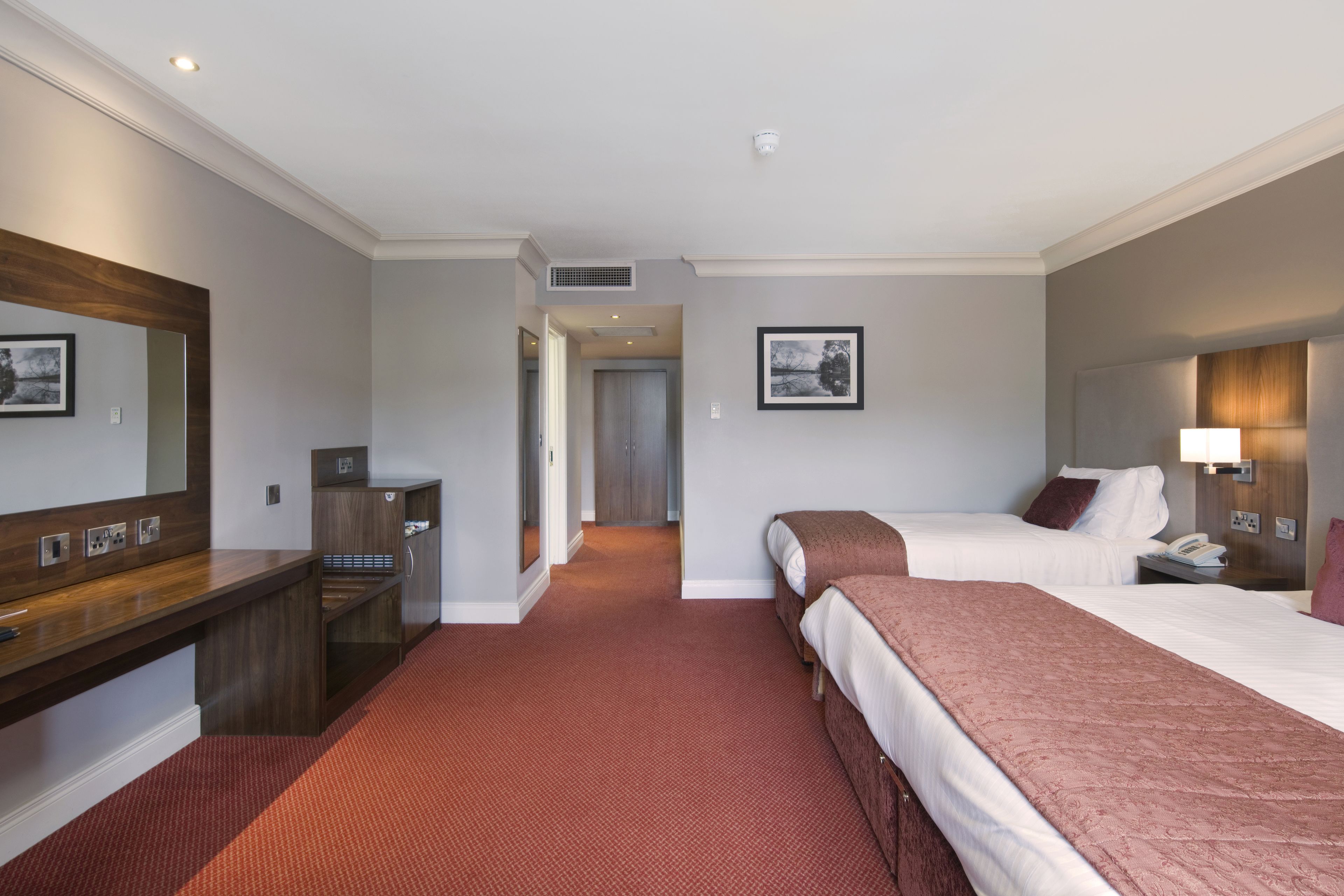A hotel room with two beds, a desk, a mirror, a bedside table, a lamp, and a framed picture on the wall.