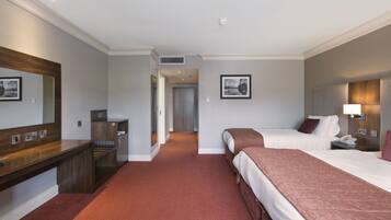 A hotel room with two beds, a desk, a mirror, a bedside table, a lamp, and a framed picture on the wall.
