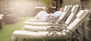 A row of sun loungers with white cushions, arranged in an outdoor patio area with a stone wall backdrop and a green artificial turf floor.