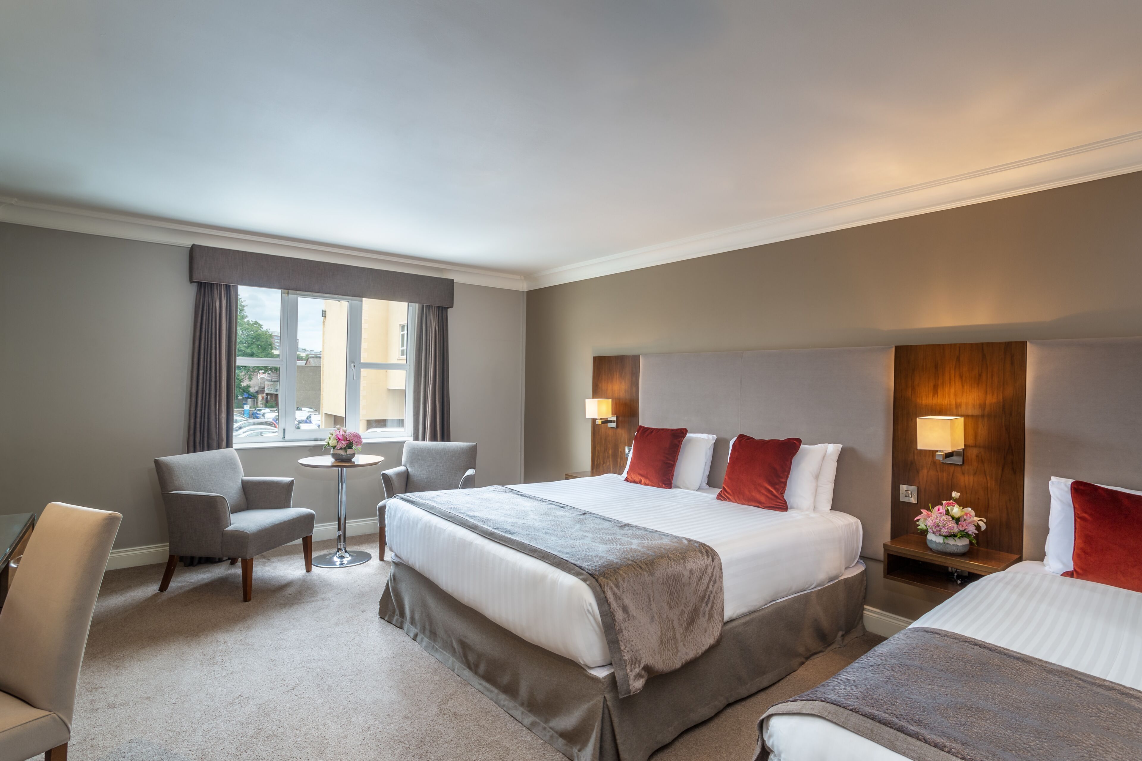 A hotel room with a large bed, a smaller bed, a desk, two chairs, a window with curtains, and a wall-mounted lamp.