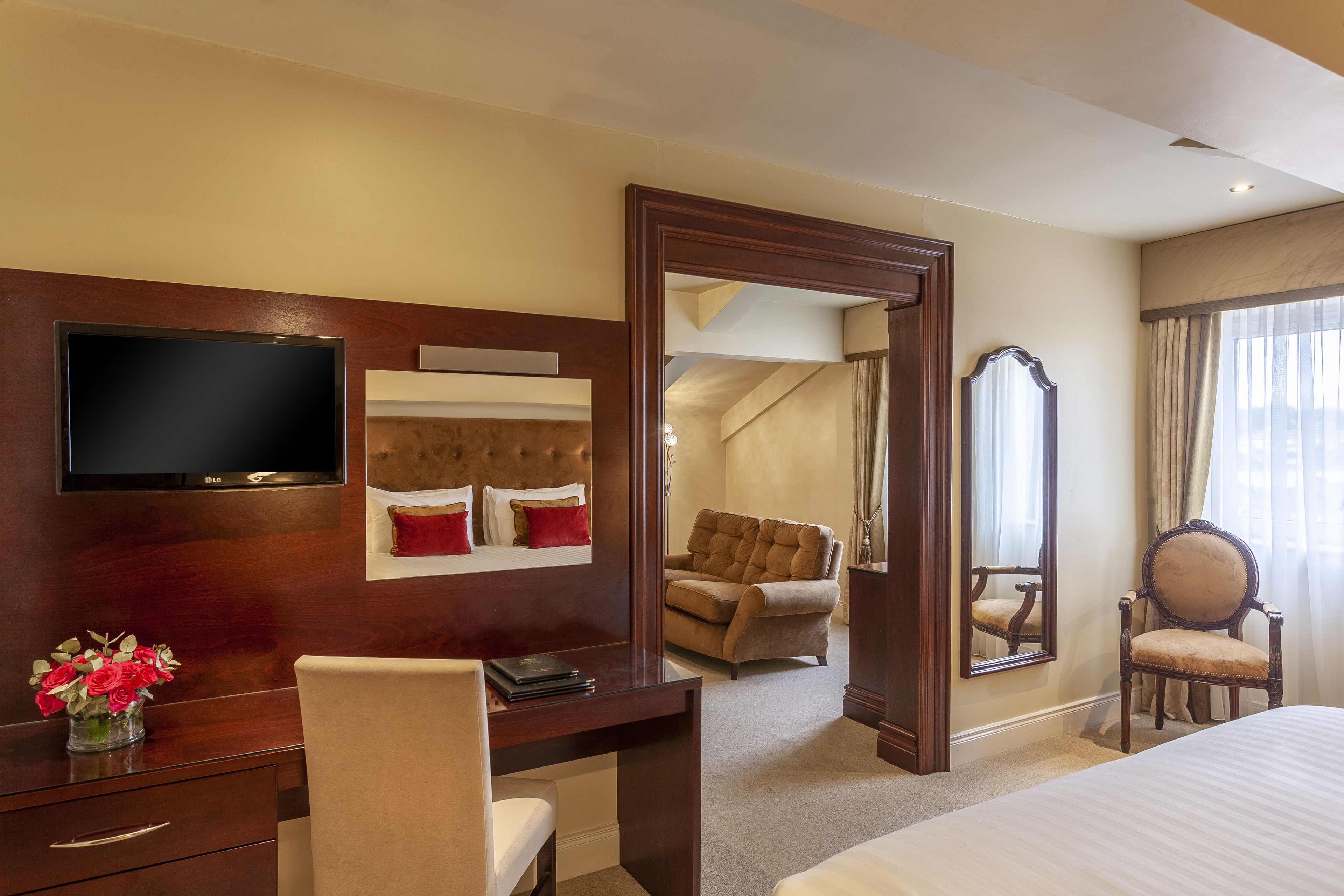 A hotel room with a bed, a desk with a TV, a chair, a mirror, and a view of another room.