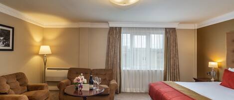 Una habitación de hotel con una cama, dos sillones, una mesita con un jarrón de flores, una lámpara y una ventana con cortinas.
