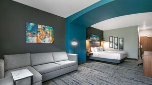 Une chambre d’hôtel avec un canapé gris, un lit, un bureau et un grand tableau abstrait accroché au mur.