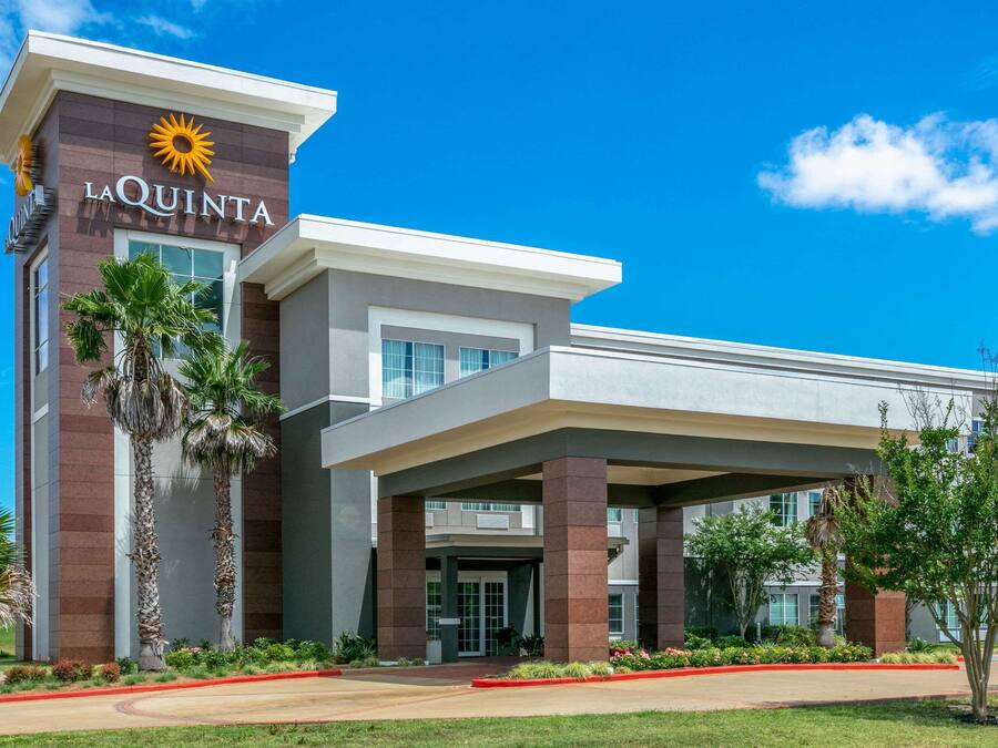 A modern hotel with a prominent 'La Quinta' sign, featuring a sun emblem. It has a covered entrance with palm trees and a well-maintained lawn.