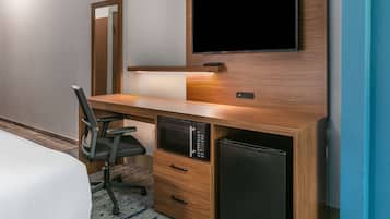 Habitación de hotel con un escritorio de madera, un televisor de pantalla plana montado encima, microondas, refrigerador, una silla y una cama con cubrecama blanco.