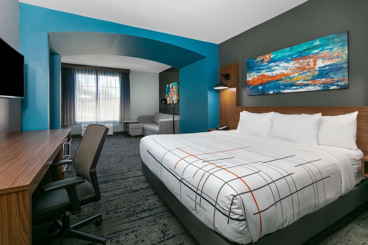 A hotel room with a bed, desk, chair, and a painting on the wall.