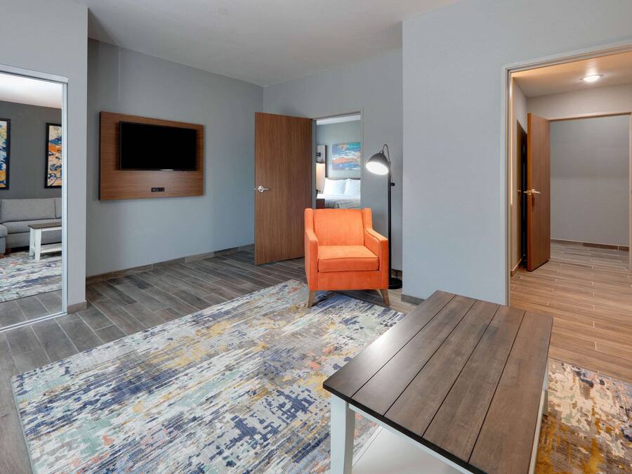 A modern hotel room with a flat-screen TV mounted on the wall, an orange armchair, a wooden table, and a colorful rug.